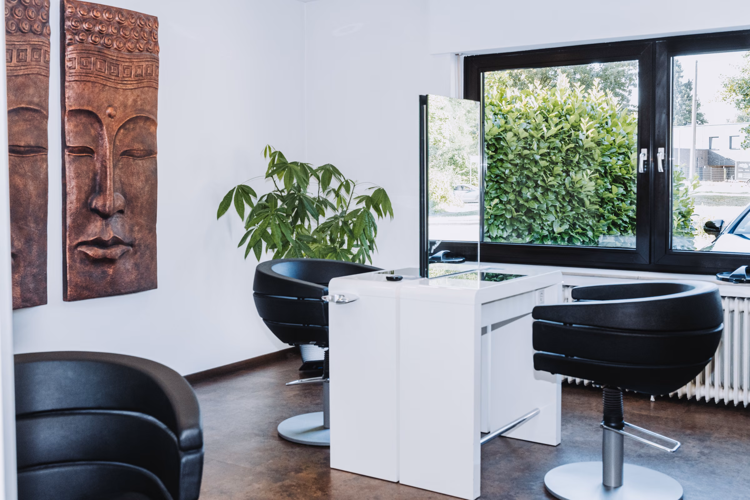 Behandlungsplatz am Fenster im Friseursalon Irena: zwei schwarze Sessel, weiße Konsole, Wandreliefs und eine Grünpflanze.