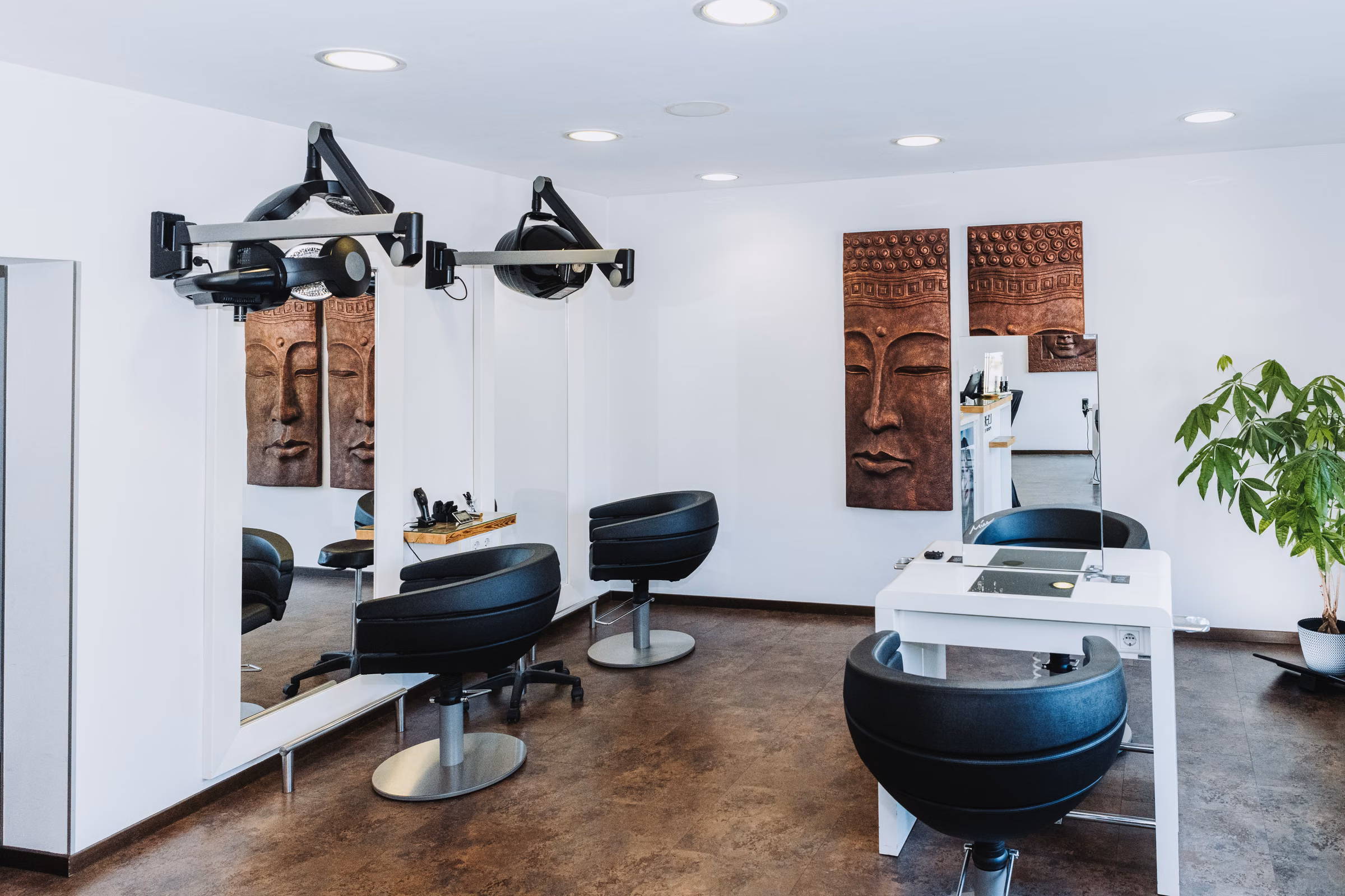 Arbeitsplatz im Friseursalon Irena: schwarzer Frisierstuhl vor Spiegel, Wandrelief und Grünpflanze, Tageslicht.